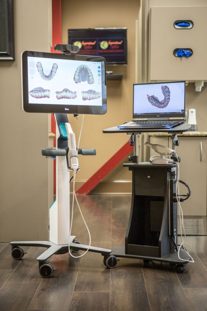 A modern dental office setup with a computer monitor displaying teeth images, a laptop, and a rolling cart with a dental mirror on top.