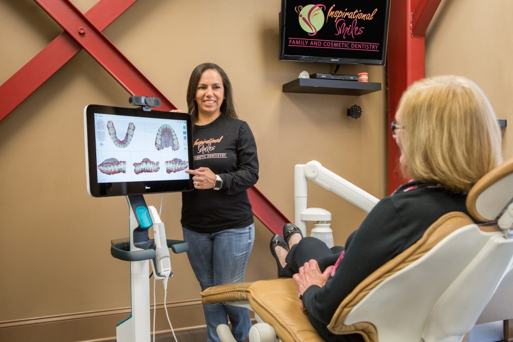 A woman stands next to a dental chair with a digital display showing teeth models, explaining dental procedures to an older woman seated in the chair.