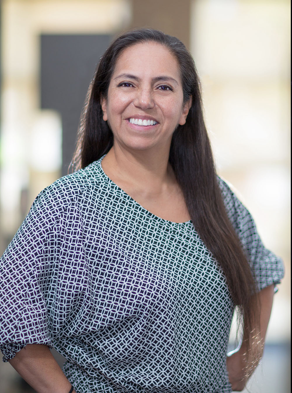 The image is a portrait of a woman standing against a blurred background. She has long hair and is wearing a patterned top, smiling slightly towards the camera.