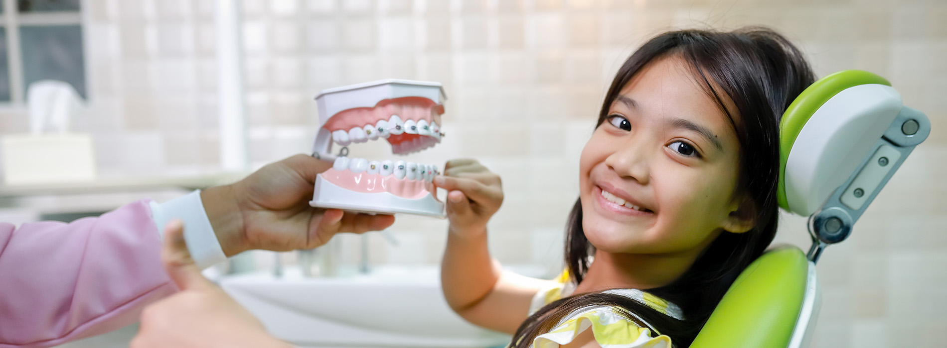 A young girl is holding up a cup of toothpaste next to an open mouth dental chair, with two people standing behind her, one holding a camera.