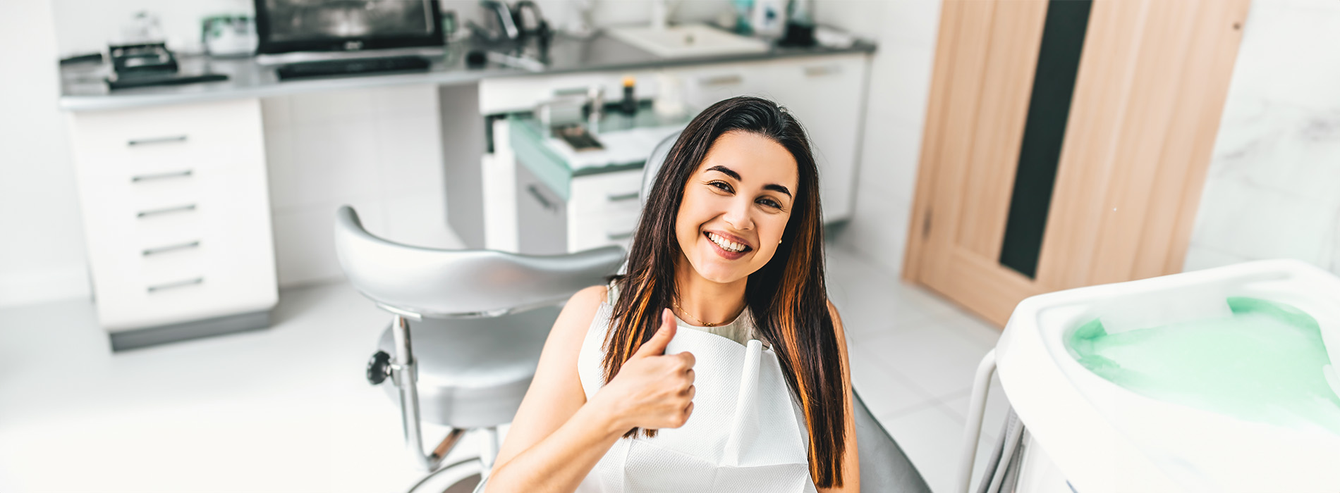 A woman posing with a thumbs-up sign in front of a dental chair, in a dental office setting.