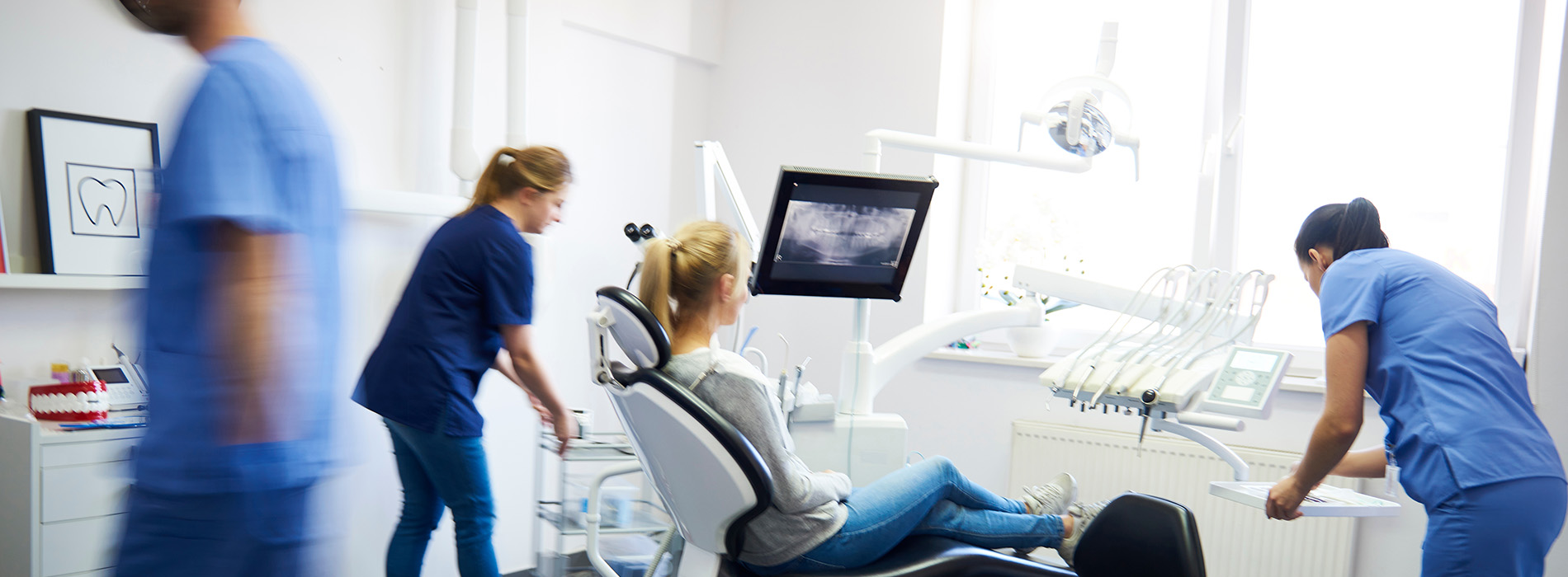 The image shows a group of healthcare professionals, likely nurses, working together in a medical setting, with one person seated at a desk with a computer screen and another standing behind her, possibly assisting or observing. They are all dressed in scrubs, suggesting they are medical personnel.