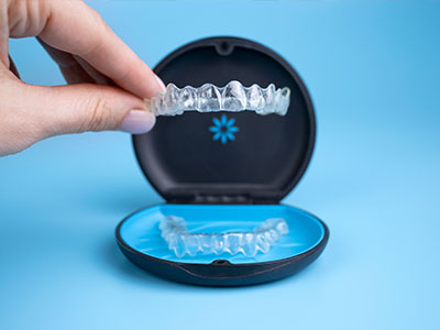 The image shows a hand holding an open case containing a set of clear dental aligners with a blue lid, against a light blue background.