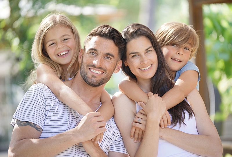 A family of four poses together on a sunny day, with a man holding two children, all smiling at the camera.