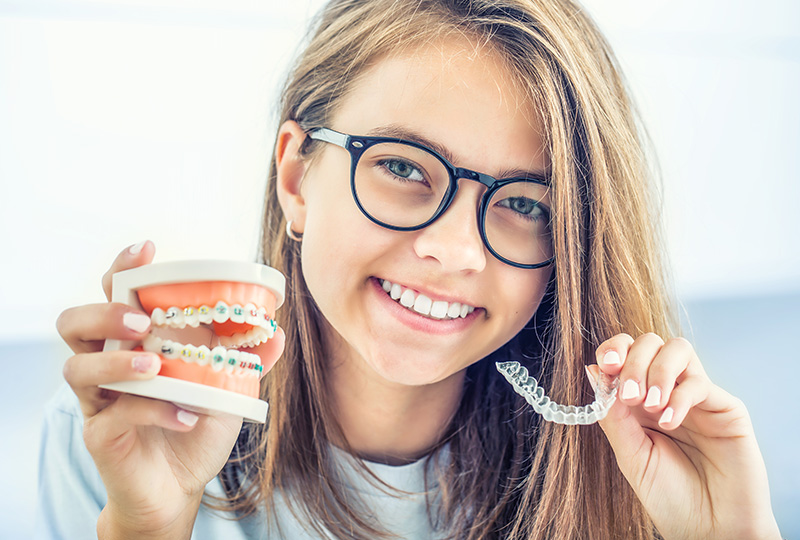 The image shows a young girl with glasses holding a toothbrush and a toothpaste tube, smiling at the camera.