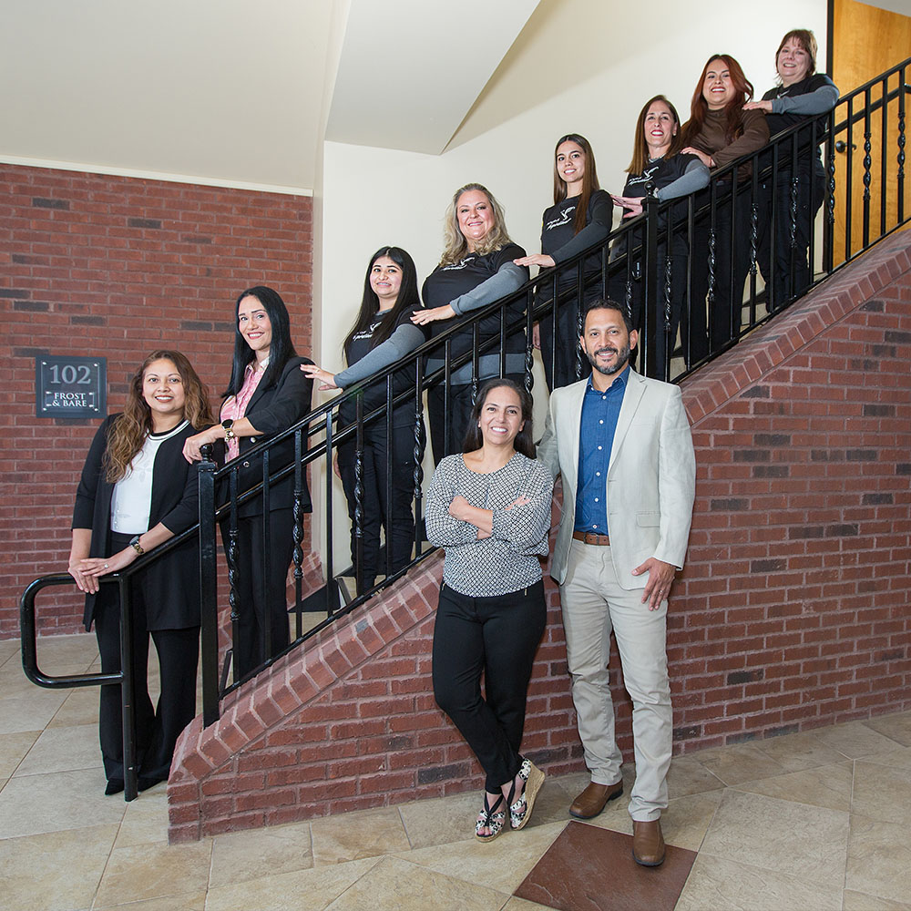 The image shows a group of individuals posing on a staircase with a handrail, including one man standing at the bottom and smiling towards the camera, and several other people standing behind him, some of whom are also smiling and looking directly at the camera, while others appear to be looking at something off-camera. The setting seems to be an indoor environment, possibly a lobby or entrance area, with a brick wall in the background and a sign that reads 101. There is no visible text providing additional context about the event or the people depicted.