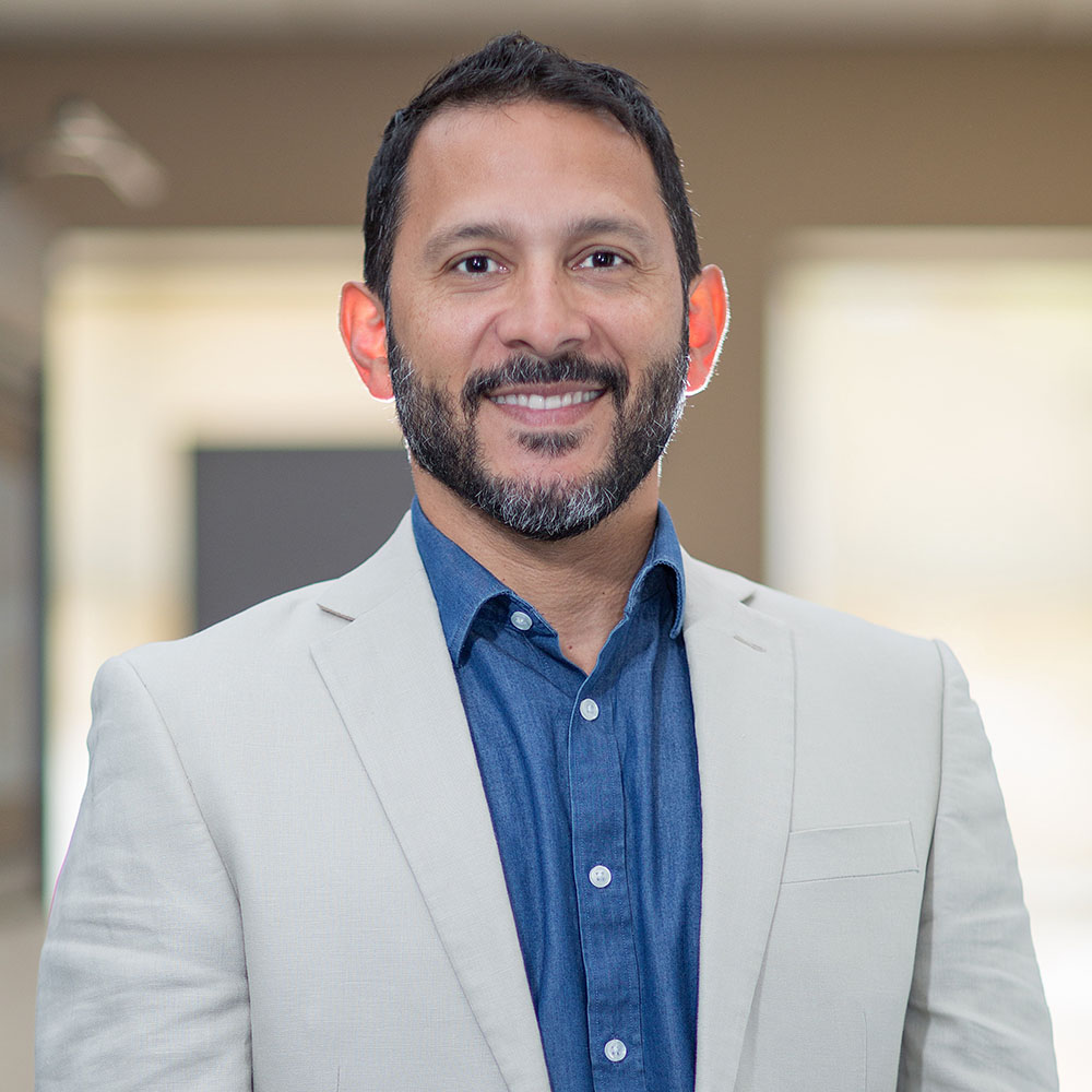 A man wearing a blue shirt stands on the left side of the frame, smiling at the camera, while another man, dressed in a white shirt and sporting a beard, smiles confidently on the right.
