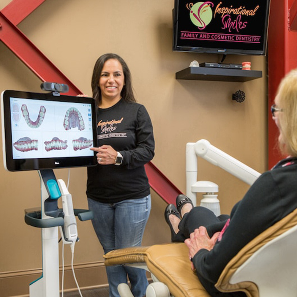 The image shows a woman standing beside a dental chair with a screen displaying a 3D model of a person s teeth, indicating a dental technology demonstration or consultation setting.