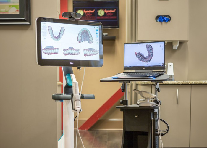 The image shows a dental office setting with equipment used for dental procedures, including a dental chair, a computer monitor displaying an X-ray, a laptop on a stand, a tablet with a mouth model, and a TV screen in the background.