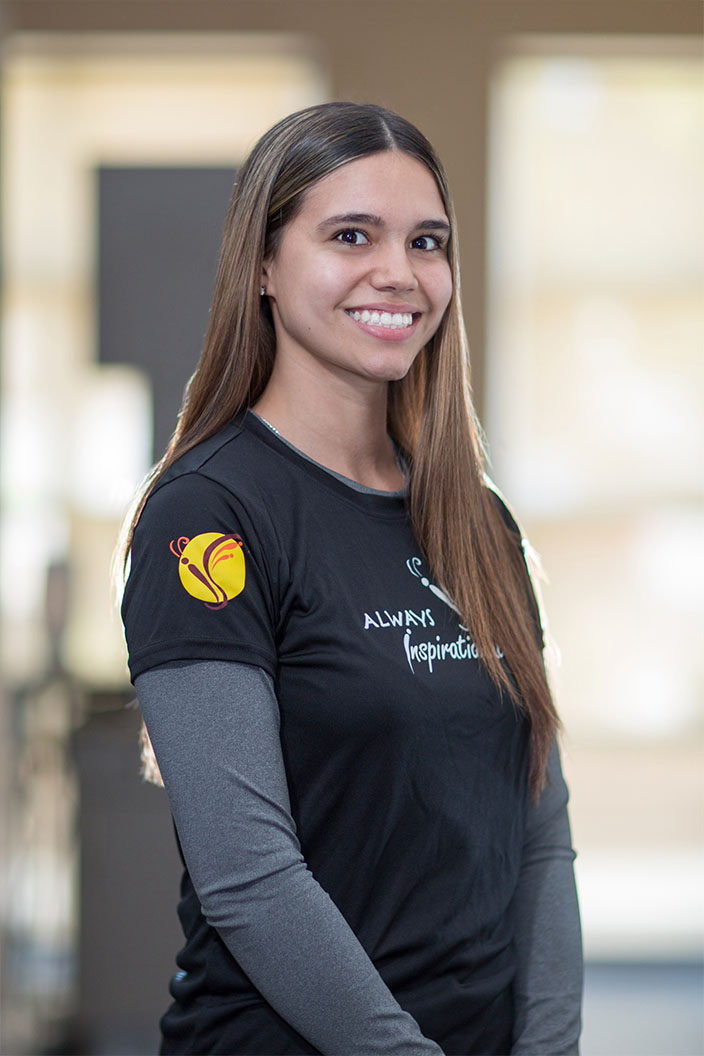 The image shows a woman standing indoors, wearing athletic attire with a logo on her shirt, smiling at the camera.