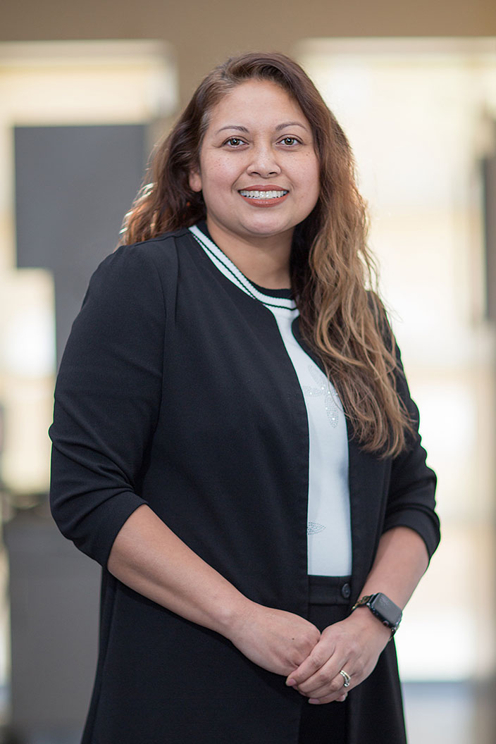 A woman with long hair, wearing a black jacket and white shirt, standing indoors with her hands together in front of her.