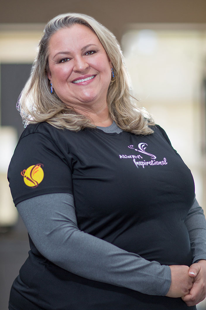 The image shows a woman wearing a black t-shirt with a logo on the front, standing indoors with her hands clasped together in front of her.