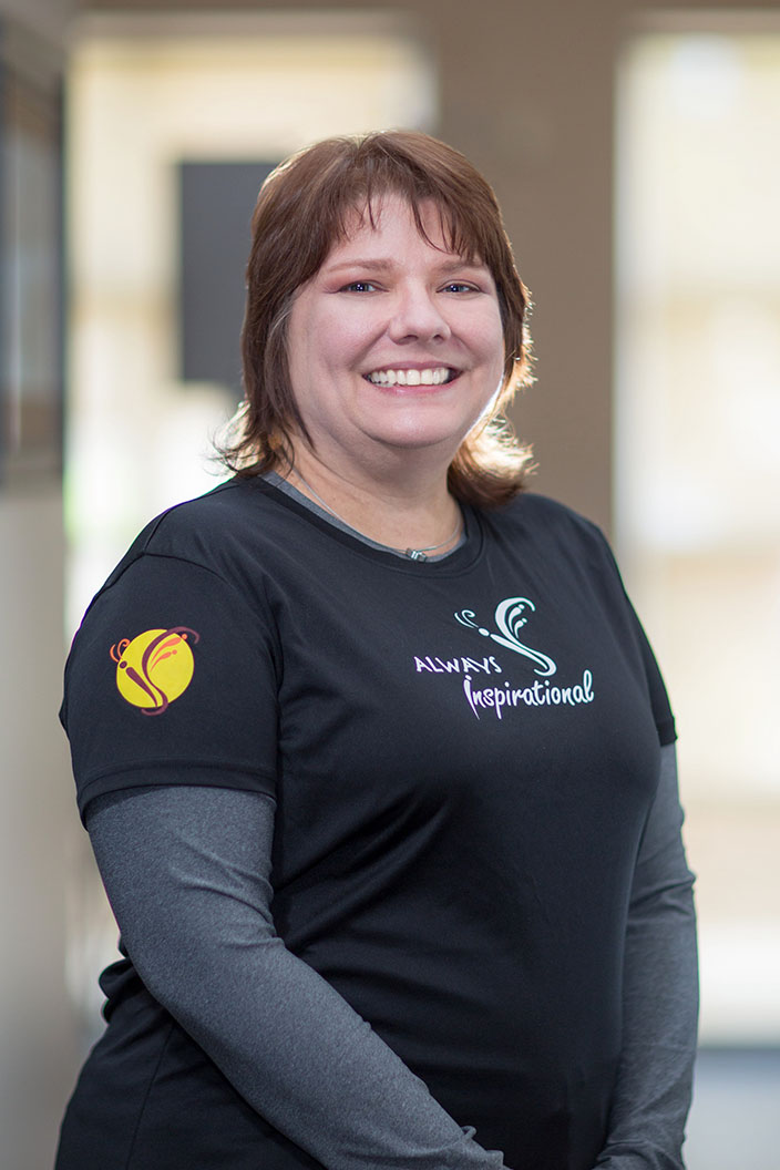 The image shows a person posing for a photo, wearing a black shirt with a logo on the front, white sleeves, and a name tag, standing indoors with a background that includes a window and blinds.