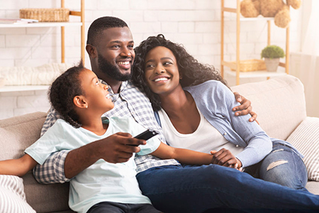 A family of three, including two adults and a child, sitting on a couch with a warm smile, watching television together.