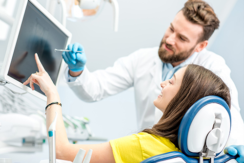 A person wearing a blue surgical mask is standing next to a woman seated in a dental chair while pointing at a computer screen, possibly explaining something related to dental technology or treatment.