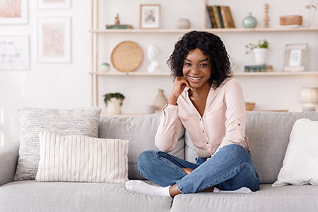 A smiling woman sitting on a couch in a living room with a white wall in the background.