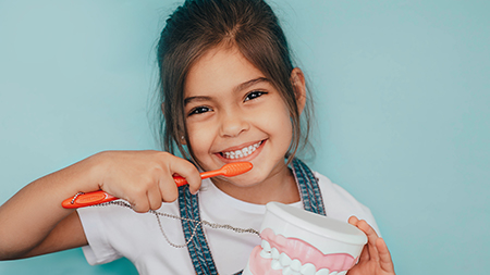 A young girl brushing her teeth with an electric toothbrush while smiling at the camera.