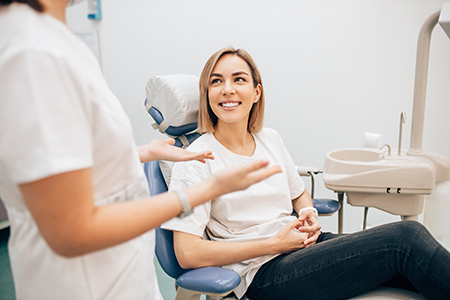 The image depicts a woman sitting in a dental chair with a smile on her face, being attended to by a dental professional who appears to be a dentist.